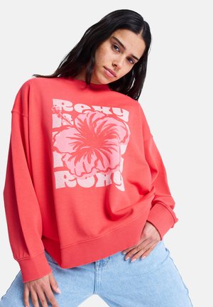 Młoda kobieta o długich ciemnych włosach ubrana w koralową bluzę z dużym kwiatem hibiskusa i napisem "Roxy" oraz jasnoniebieskie dżinsy, pozująca.