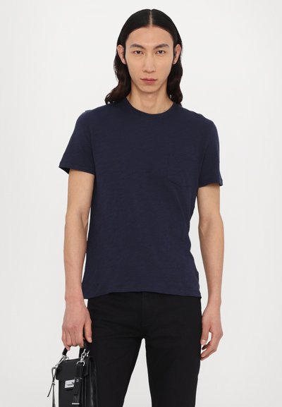 Camiseta de manga corta azul marino con cuello redondo y un bolsillo en el pecho, tejido texturizado, combinada con pantalones negros y un bolso pequeño negro.