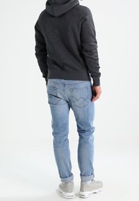 Hombre vestido con sudadera gris oscuro, jeans azul claro arremangados y zapatillas blancas, de pie con la espalda hacia la cámara sobre un fondo blanco.