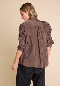 Brun, satengbluse med puffede ærmer, høj krave og knapdetaljer ved manchetterne; glat tekstur; rygplade design.