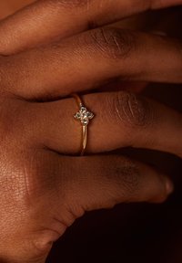 Gouden ring met een bloemmotief, met vier kleine heldere edelstenen in het midden, getoond op een gestructureerde bruine hand.