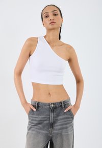 White ribbed crop top features an asymmetrical design with one shoulder. Paired with high-waisted, faded gray denim jeans.