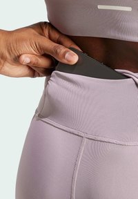 Leggings sportivi di un delicato viola chiaro con un alto elastico in vita, dotati di una tasca laterale e una texture di tessuto liscia. Una mano sta infilando un telefono nella tasca.