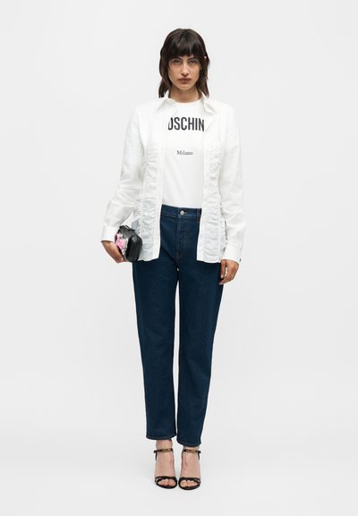 Γυναίκα που φοράει λευκό πουκάμισο με βολάν πάνω από λευκό t-shirt Moschino Milano, σκουρομπλε τζιν, μαύρα ψηλοτάκουνα με λουράκια και κρατάει μαύρο φάκελο.