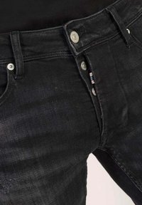 Primer plano de unos jeans de mezclilla negra con cierre de botones, remaches de metal, trabillas para cinturón y una pequeña etiqueta de tela a rayas en la costura del cierre.