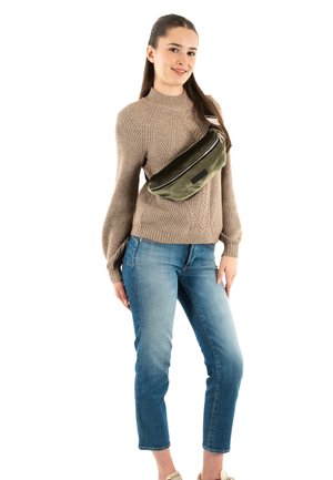 Riñonera verde oliva con cremallera brillante, llevada cruzada sobre un suéter beige y unos vaqueros azules. La bolsa tiene una textura suave y un diseño minimalista.