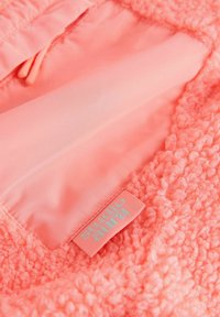 Korall rózsaszín polárdzseki belső rész sima béléssel. Texturált külső felülettel és világoskék "studio aura" feliratú cimkével.