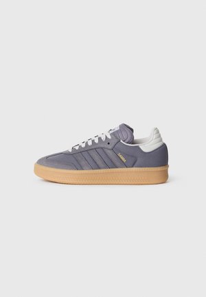 Szary but sportowy Adidas Samba z gumową podeszwą, białymi sznurowadłami i białą zakładką na pięcie, pokazany z profilu bocznego na jednolitym tle.