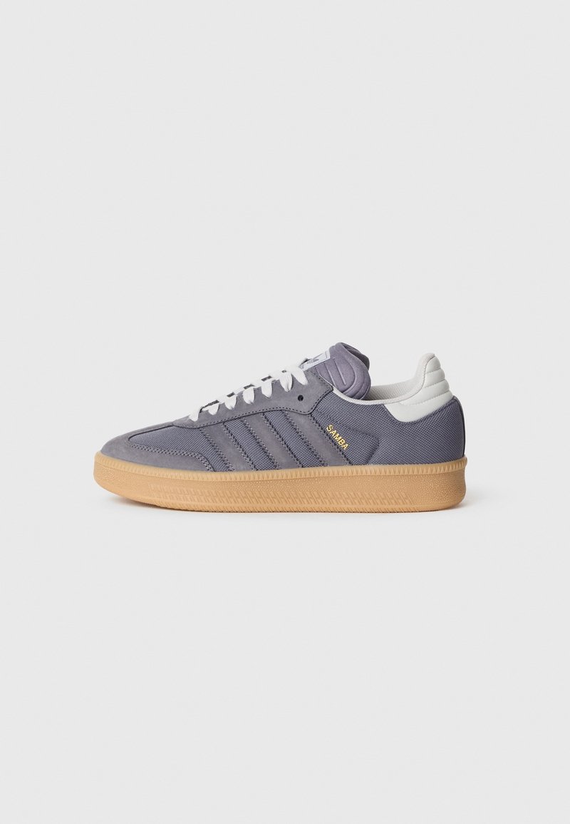 Szary but sportowy Adidas Samba z gumową podeszwą, białymi sznurowadłami i białą zakładką na pięcie, pokazany z profilu bocznego na jednolitym tle.