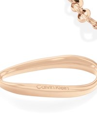 Rose gull metallarmbånd med en avlang oval form, med en jevn tekstur og preget "Calvin Klein"-logo på siden.