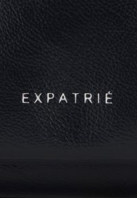 Schwarze, strukturierte Lederoberfläche mit prägnant geprägt silberner Schrift "EXPATRIÉ". Minimalistisches Design ohne zusätzliche Akzente.