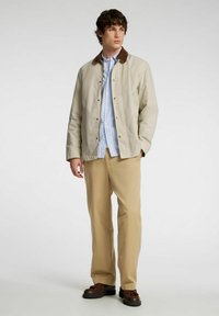 Beige sako s hnědým límcem, se zapínáním na knoflíky a postranními kapsami, nošené přes modro-pruhovanou košili a béžové kalhoty, s hnědými botami.