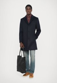 Marineblauer Wollmantel, vorne zugeknöpft, kombiniert mit einem kastanienbraunen Hemd. Helle blaue Jeans und braune Sneakers, hält eine schwarze Tragetasche.