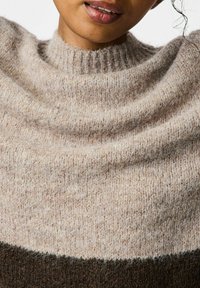 Pull en tricot de couleur beige clair avec un col haut texturé et côtelé. Présente un tissu superposé avec une stripe marron foncé au niveau de l'ourlet.