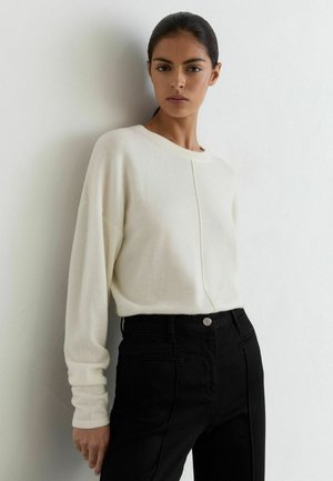 Kvinde med mørkt hår samlet bagpå, iført en hvid langærmet sweater og sorte bukser, står op ad en hvid væg.