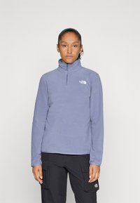 Cilvēks, kas valkā lavandas krāsas puszipa flīsi ar "The North Face" logotipu un melnām kabatas biksēm, stāvot pret viegli pelēku fonu.