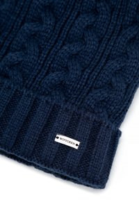 Tmavě modrá pletená čepice s tlustým žebrovaným lemem, zdobená texturovaným copánkovým vzorem. Na lemu je stříbrná štítek s nápisem „WITTCHEN“.