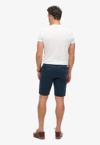 Marineblauwe shorts van katoen. Voorzien van achterzakken met knopen en een opgerolde zoom. Gecombineerd met een effen wit T-shirt en bruine schoenen.