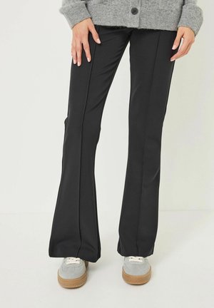 Pantalons évasés noirs en tissu texturé ; offrent une coupe lisse au niveau des cuisses et un large ourlet. Associés à des baskets grises.