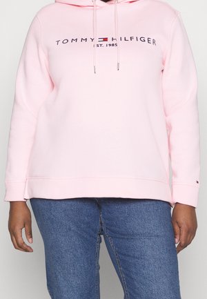 Osoba ubrana w jasnoróżową bluzę z kapturem Tommy Hilfiger z logo i napisem "EST. 1985", w połączeniu z niebieskimi dżinsami, stojąca na białym tle.