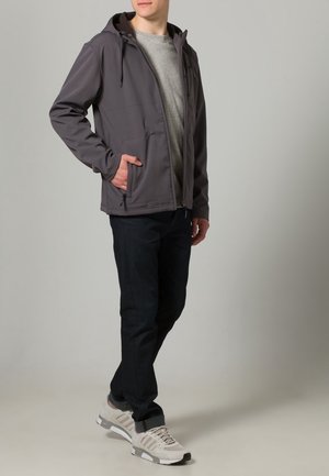 Hombre vestido con una chaqueta gris con capucha sobre una camiseta gris claro, pantalones negros y zapatillas gris claro, de pie con una mano en el bolsillo de la chaqueta.