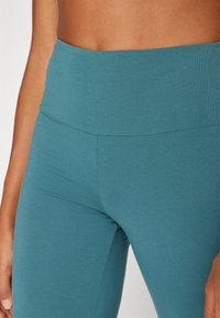 Teal színű, magas derekú, rugalmas anyagból készült leggings. Simaság jellemzi, testhez álló kialakítással, széles derékpánttal és láthatatlan varrásokkal.