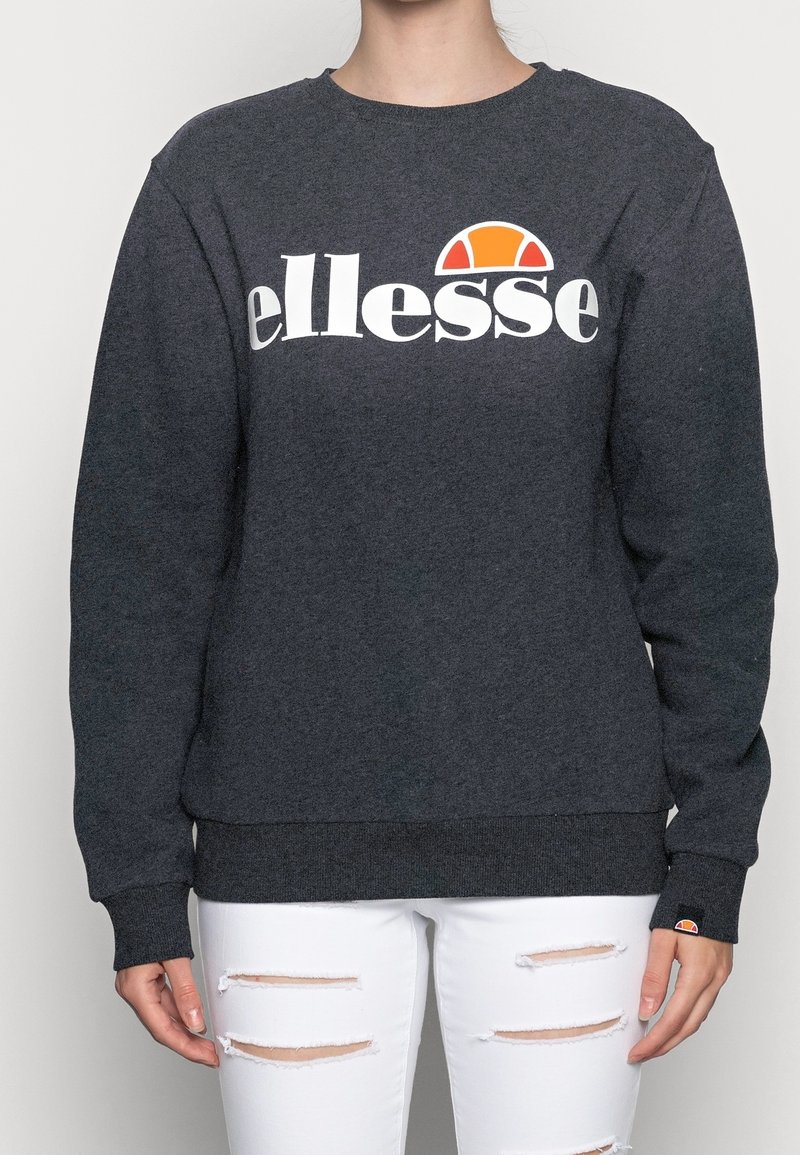 Personne portant un sweat-shirt ellesse gris foncé avec un logo orange et blanc, associé à un jean blanc déchiré, debout devant un fond uni.