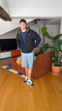 Joven con chaqueta azul marino y pantalones cortos de mezclilla sonriendo en una sala de estar con un sofá marrón, una planta verde grande, un televisor y suelo de madera.