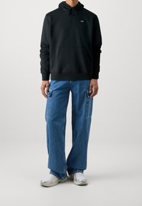 Persona che indossa un felpa nera con cappuccio, jeans cargo blu larghi con tasche a patta e sneakers bianche, in piedi su uno sfondo chiaro e uniforme.