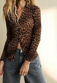 Camisa de botones con estampado de leopardo y textura acanalada, en colores marrón y negro, con cuello en v. Combinada con jeans de talle alto en azul desteñido.