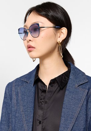 Giovane donna con capelli neri che indossa occhiali da sole sfumati blu, orecchini a catena d'oro, camicia nera e blazer di jeans blu, guarda di lato.