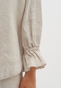Top in lino beige con polsini arricciati e svasati; texture morbida; dettaglio dell'orlo a strati; vista ravvicinata della manica e del polso.