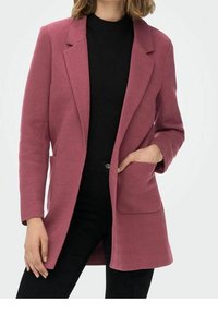 Blazer rose à chevrons avec un col cranté, une fermeture à un bouton, deux poches avant et une coupe ajustée, porté sur un haut noir.