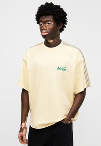 Κίτρινο βαμβακερό T-shirt με κοντά μανίκια, διαθέτει πράσινο λογότυπο Adidas στο στήθος και τρεις γκρι ρίγες στους ώμους, σε άνετη γραμμή.