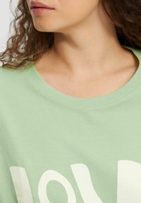 Jasnozielona bawełniana koszulka z okrągłym dekoltem, z wyraźnym białym napisem na piersi. Gładka faktura, casualowy design.