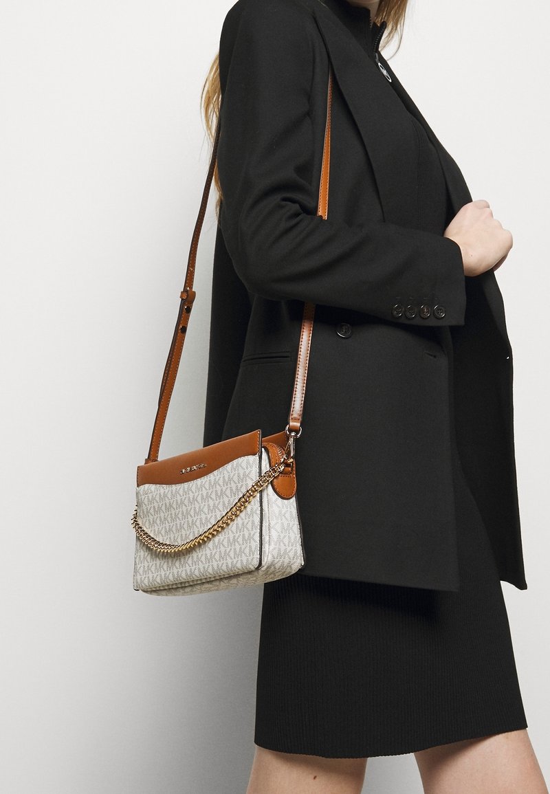 Femme portant un blazer noir et une jupe, porte un sac bandoulière beige et marron avec un détail chaîne dorée et un motif logo répété.