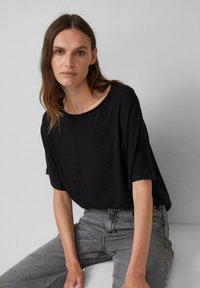 Kobieta z długimi brązowymi włosami ma na sobie czarną bluzkę z krótkim rękawem i szare dżinsy, siedzi na tle jasnoszarym.