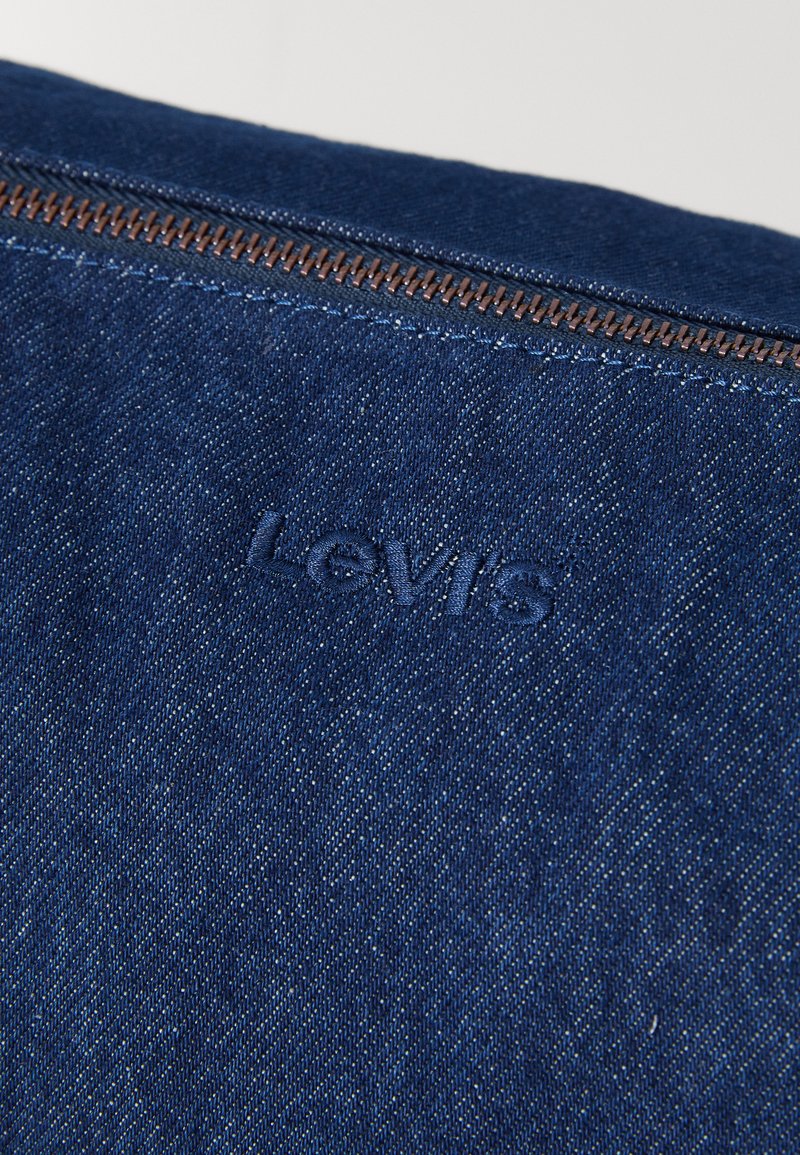 Levi's denimstof i mørkeblå med en struktureret overflade, udstyret med en brun metallynlås og prægede logodetaljer.