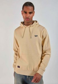 Homme en sweat à capuche beige avec poche frontale et logo noir "Redskins", portant un jean bleu, se tenant devant un fond clair.