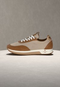 Ανοιχτό καφέ και μπεζ αθλητικό παπούτσι με λευκά κορδόνια και λευκή σόλα, τοποθετημένο σε ανοιχτή επιφάνεια με γκρι φόντο.