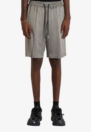 Uomo che indossa pantaloncini grigi larghi con coulisse nere, calzini neri alla caviglia e scarpe da ginnastica nere, in piedi su uno sfondo bianco.