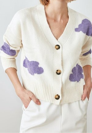 Cardigan crème avec motifs de nuages violets, grands boutons et ourlet côtelé, porté sur un pantalon blanc avec les mains dans les poches.