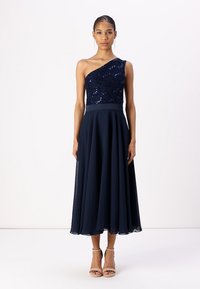 Robe de soirée bleu marine avec une épaule, corsage à paillettes et jupe en chiffon fluide. Des escarpins beiges à bout ouvert complètent le look.
