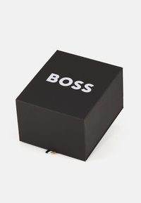 Boîte rectangulaire noire avec une finition lisse, comportant le texte blanc "BOSS" sur le dessus et une étiquette décorative en noir, blanc et or au bas.