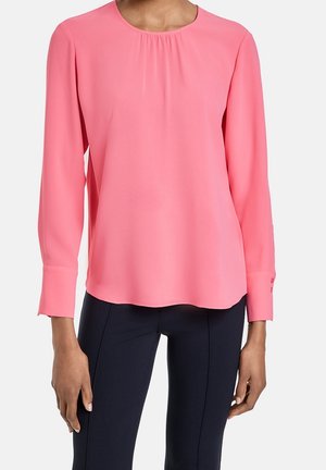 Femme portant un chemisier rose à manches longues avec un col rond et un pantalon noir ajusté sur un fond blanc.
