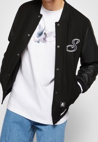 Persoon in een zwarte varsityjas met leren mouwen en een patch met de letter "S", over een wit shirt met een afbeelding van twee duiven, en blauwe jeans.