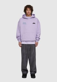 Felpa viola con testo "Retro Future", tasca frontale e logo nero; abbinata a pantaloni della tuta grigio scuro e scarpe nere.