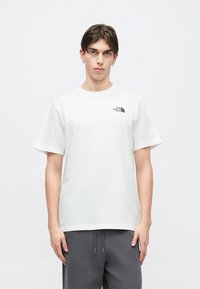 Λευκό βαμβακερό μπλουζάκι με κοντά μανίκια, στρογγυλή λαιμόκοψη και μαύρο λογότυπο στο αριστερό στήθος. Φοριέται με σκούρο γκρι παντελόνι.