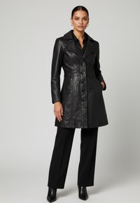 Kvinna klädd i en svart knäppt skinnrock, svarta byxor och svarta ankelstövlar, stående med ena handen i rockfickan mot en grå bakgrund.