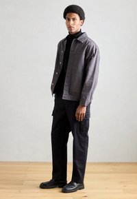 Chaqueta gris oscuro para hombre con botones en la parte delantera, combinada con un cuello de tortuga negro, pantalones cargo oscuros y zapatos negros de slip-on sobre un suelo de madera.